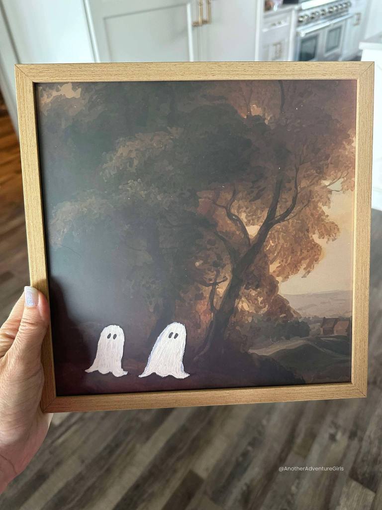 woman holding a square painting of a tree in the fall with two white ghosts added to the bottom left corner under the tree