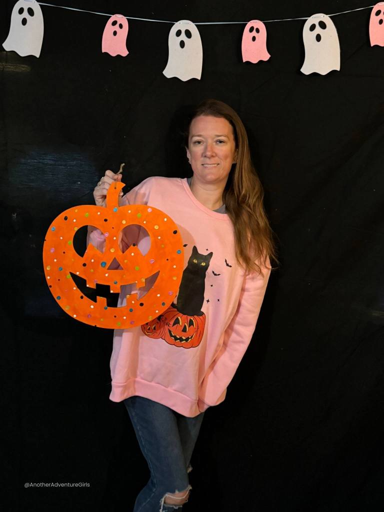 tiffany standing in front of a black and pink halloween background holding a wooden pumpkin painted with orange and colorful polka dots