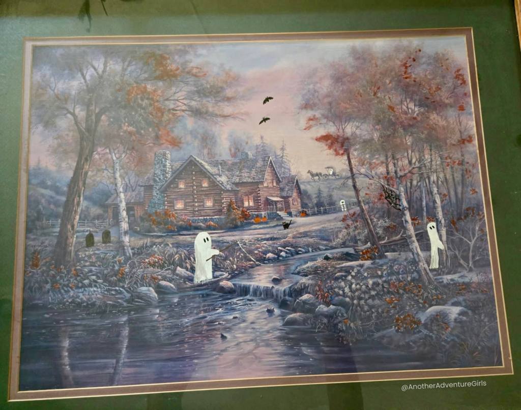 painting with a house in the woods behind a stream of water in the fall with the addition of tiny ghosts and little Halloween details added