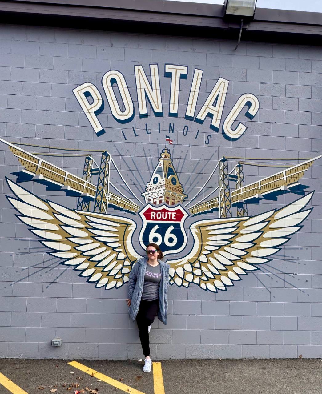 Tiffany posing in front of a mural with large wings with a route 66 sign in the center and Pontiac Illinois text overhead